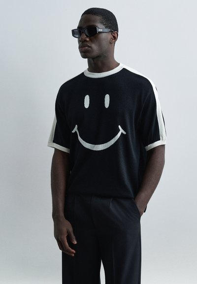 Hombre con camisa de manga corta en blanco y negro con un gran gráfico de cara sonriente, pantalones negros y gafas de sol negras rectangulares, de pie contra un fondo claro.