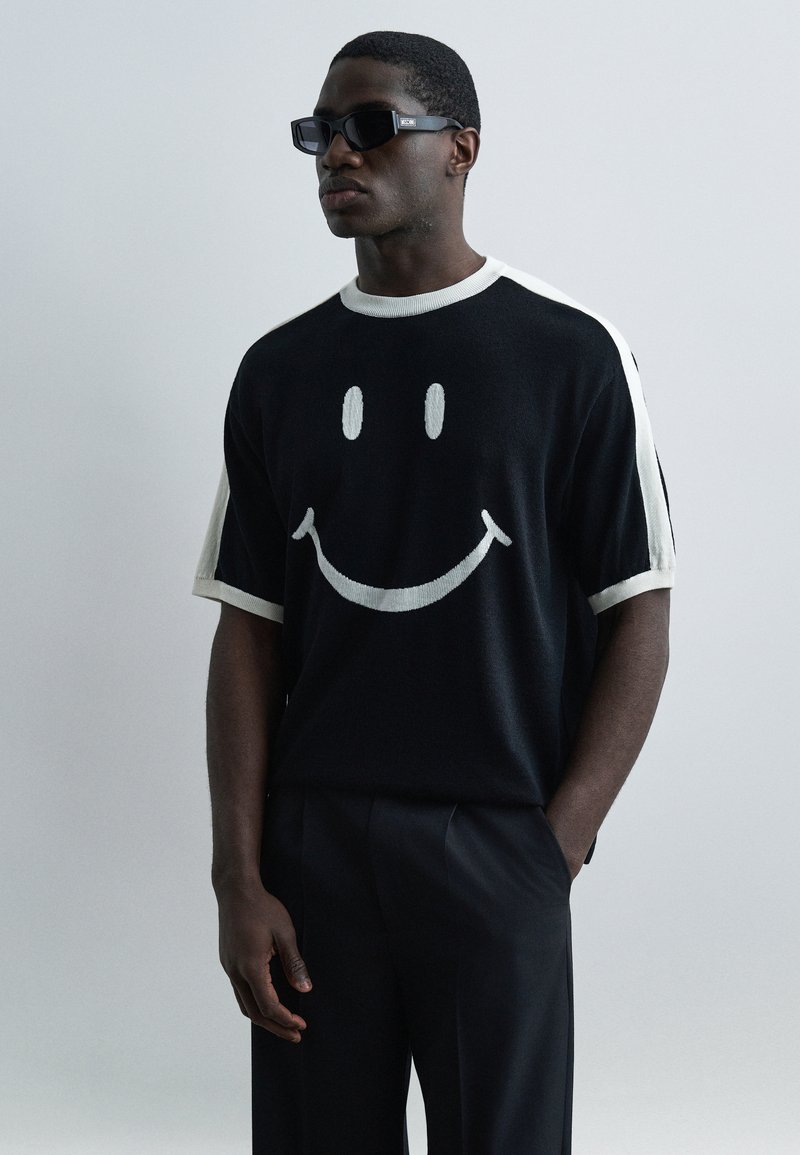 Άνδρας που φοράει μαύρο και λευκό κοντομάνικο πουκάμισο με μεγάλο γραφικό χαμογελαστό πρόσωπο, μαύρο παντελόνι και ορθογώνια μαύρα γυαλιά ηλίου, στέκεται μπροστά σε ανοιχτόχρωμο φόντο.