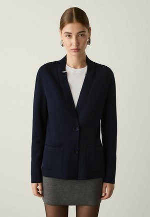 Frau mit zurückgekämmtem Haar, die eine dunkelblaue Strickjacke über einem weißen Oberteil und einem grauen Rock trägt, steht vor einem einfarbigen hellen Hintergrund.