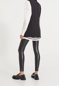 Czarna bezrękawnikowa kamizelka na tle fakturowanego, białego swetra z dzianiny, w połączeniu z czarnymi legginsami z sztucznej skóry oraz błyszczącymi, czarnymi loafersami na grubych podeszwach.