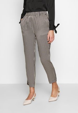 Frau trägt schwarz-weiß karierte, knöchellange Hosen mit Taschen, eine schwarze Bluse mit langen Ärmeln und beige niedrig-absätzige Slingback-Schuhe.