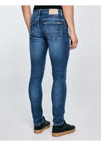 Vīrietis, kurš valkā šauras zila džinsa bikses un melnus sporta apavus ar brūnām zolēm, stāv pret vienkāršu fonu.