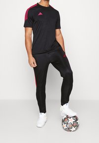 Svart sportt-shirt och byxor med rosa accenter, med Adidas-loggor. Modellen står på en fotboll, klädd i vita sportskor.