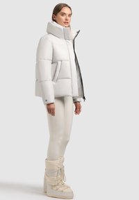 Hvid pufferjakke med høj krave, firkantet form og store quiltede sektioner. Sammen med cremefarvede leggings og vinterstøvler.