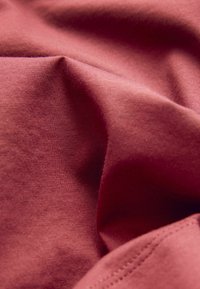 Weiches, zerknittertes, rötlich-braunes Gewebe mit sichtbarer Webstruktur und genähtem Naht in der unteren rechten Ecke.