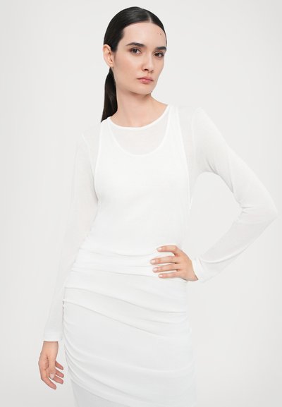 Witte lange mouw top met een ronde halslijn, gelaagde rimpeling bij de taille en een gladde, lichte stof. Model poseert met de hand op de heup.