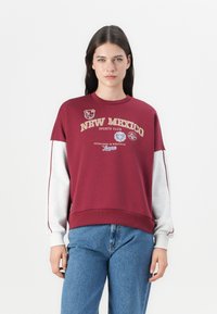 Bordeaux sweatshirt met grijze mouwen, voorzien van de tekst "NEW MEXICO SPORTS CLUB" en patches. Klassieke ronde hals en een ontspannen pasvorm.