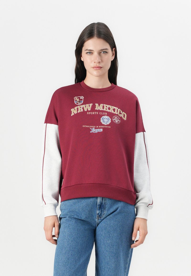 Sweatshirt bourgogne avec des manches grises, affichant le texte "CLUB DE SPORT DU NOUVEAU MEXIQUE" et des écussons. Col ras du cou classique et coupe décontractée.