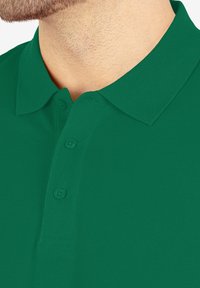 Groene polo met een gestructureerde stof, voorzien van een kraag en een drie-knopen sluiting. Close-up toont het halsgebied en de details van het shirt.