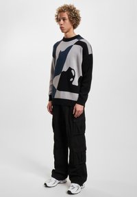 Knitted sweater featuring geometric patterns in gray, black, and navy; crew neck design with ribbed edges; worn with black cargo pants.
