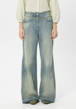 Frau trägt helle, ausgewaschene Jeans mit weitem Bein, doppelt geknöpftem Verschluss und ausgefranstem Bund über grünen, mit Nieten besetzten Schuhen.