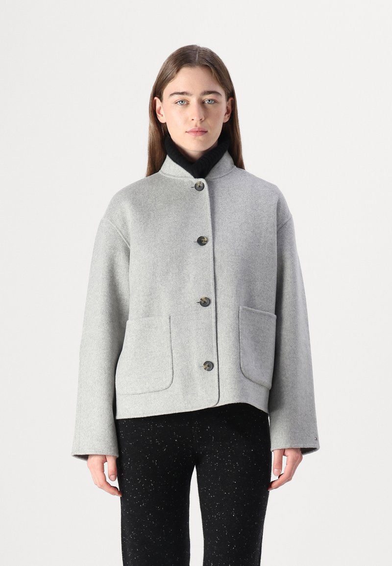 Grå uldjakke med ståkrave, frontknaplukning og to sidelommer. Struktureret stof, cropped design, parret med sorte bukser.