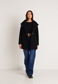 Čierny kabát z vlnenej zmesi s veľkým golierom a dvojradovým zapínaním, spárovaný s čiernym topom a modrými džínsami s vysokým pásom.