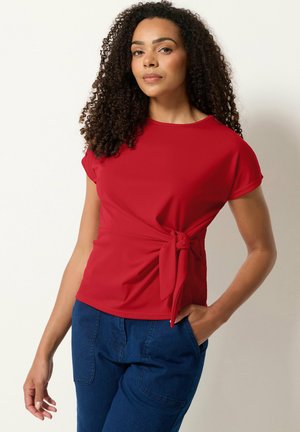 Donna con capelli ricci che indossa una maglietta rossa a maniche corte con fiocco laterale e jeans blu, in piedi con una mano in tasca su sfondo semplice.