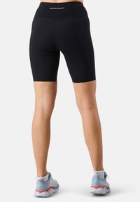 Shorts da ciclismo neri a vita alta con una texture liscia e un dettaglio con logo sulla cintura, abbinati a scarpe da ginnastica azzurro chiaro.