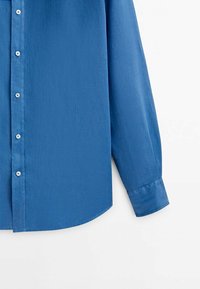 Blå skjorta med knäppning, tillverkad av slätt tyg med krage, långa ärmar och rundad nederkant. Har vita knappar fram.