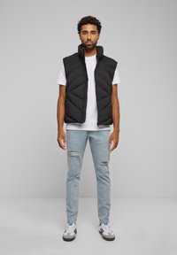 Schwarze Steppweste mit hohem Kragen und gestepptem Design. Getragen über einem weißen T-Shirt, kombiniert mit hellen, distressed Jeans und Sneakers.