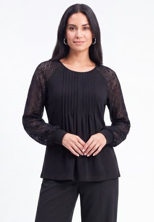 Femme portant un chemisier noir avec des manches longues en dentelle et un devant plissé, debout, les mains doucement jointes devant un fond blanc.