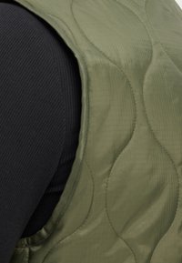 Gilet verde oliva trapuntato realizzato in tessuto liscio, con un motivo arrotondato. Sottostrato nero a costine parzialmente visibile sotto.