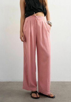 Woman wearing loose pink wide-leg pants tied at the waist, black sleeveless top, black sandals, and chunky silver bracelet, standing indoors.