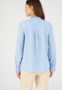 Blusa de manga larga azul claro con cuello con volantes. Presenta puños abotonados y un pliegue en la parte posterior, hecha de una tela suave.