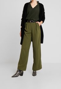 Cardigan nero, blouse verde testurizzato, pantaloni larghi oliva, cintura nera e stivali alla caviglia in stampa serpente con tacco quadrato.