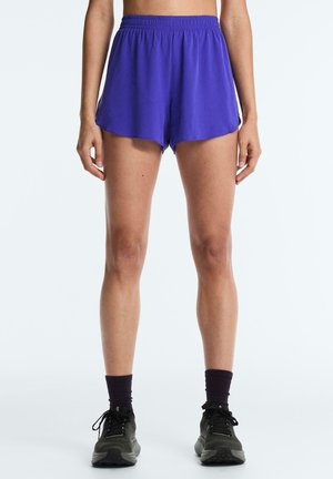 Femeie purtând pantaloni scurți sportivi de culoare violet aprins, șosete negre până la glezne și pantofi de alergare negri, stând cu brațele relaxate pe lângă corp.