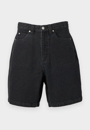 Mand iført sorte denimshorts, sorte sneakers med hvide snørebånd, hvide sokker og en sort t-shirt, stående på en hvid baggrund.
