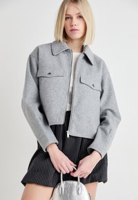 Hallikas lühike jakk, mis on valmistatud tekstuuriga kangast, millel on kraega kaelus, kaks esist pocketi ja lukuga sulgemine. Rikka hõbedaste detailidega.