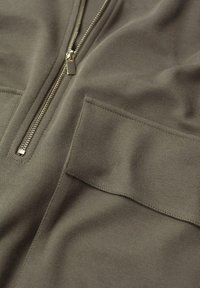 Prenda de color verde oliva con cremallera, fabricada en un tejido texturizado, que cuenta con una cremallera prominente y un bolsillo cuadrado en la parte frontal.