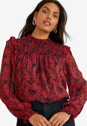 Paita dengan cetakan ular merah dan hitam, dilengkapi dengan kerah ruffle tinggi, bodice beralun, dan lengan panjang dengan manset yang berkumpul. Kainnya lembut dan transparan.