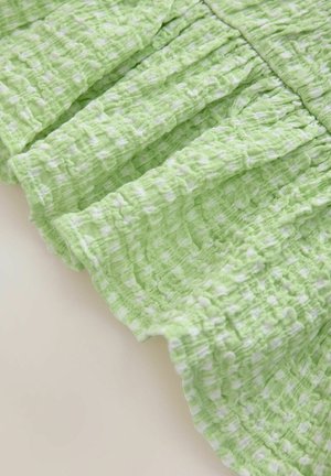 Grüner, texturierteter Stoff mit einem zerknitterten Muster und weißen Akzenten, der Rüschen entlang des Randes aufweist. Das Material scheint leicht und atmungsaktiv zu sein.