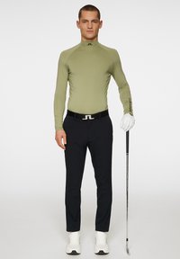 Långärmad, olivgrön sporttröja med hög krage, kombinerad med svarta skräddarsydda byxor och vita sneakers, hållande en golfklubba i ena handen.