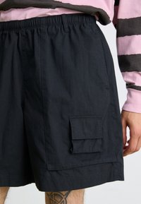 Black shorts made of lightweight fabric, featuring an elastic waistband, side pocket, and a flap pocket on the thigh. Smooth texture.