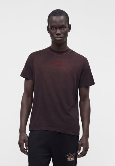 Homme portant un t-shirt sombre avec un petit texte rouge sur la poitrine et un pantalon noir avec un motif coloré près de la poche, debout devant un fond blanc.