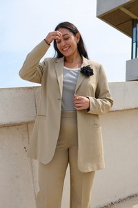 Beige sako s volnějším střihem, se dvěma předními kapsami, nošené přes pruhované tričko. Má texturovanou černou květinovou dekoraci na klopě.