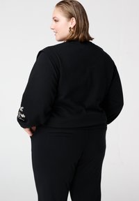 Zwarte cropped jas met lange mouwen en bloemrijke borduursels op de polsen. Gemaakt van zachte stof, gecombineerd met op maat gemaakte zwarte broek.