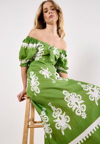 Grünes schulterfreies Kleid mit weißen Blumenmustern, gepufften Ärmeln und fließender Silhouette; der strukturierte Stoff erhöht das visuelle Interesse.