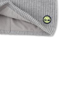 Bonnet en tricot gris clair avec doublure en polaire douce, présentant un petit patch logo circulaire avec un motif d'arbre en jaune et bleu marine.