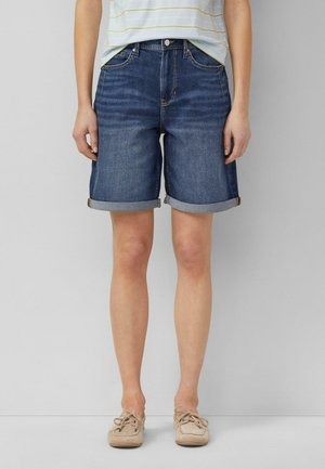 Personne portant un short en jean bleu foncé retroussé, une chemise claire rayée et des chaussures beiges à lacets, debout devant un fond gris uni.
