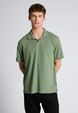 Młody mężczyzna z kręconymi włosami, ubrany w jasnozieloną koszulkę polo z krótkim rękawem i czarne spodnie, stojący na jednolitym jasnym tle.