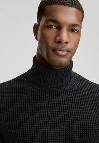 Pull en maille noir avec col roulé, présentant un motif texturé et un col côtelé. La vue en gros plan se concentre sur le décolleté et l'ajustement.