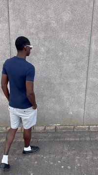 Camisa de manga corta azul marino, pantalones cortos grises claros, calcetines blancos y zapatos negros; de pie contra una pared gris texturizada.
