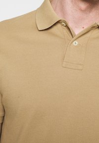 Béžové polo tričko vyrobené z texturované bavlny, s klasickým límcem, zapínáním na dva knoflíky a jemným tkaným vzorem.