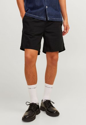 Hombre con pantalones cortos negros, calcetines blancos con texto, zapatos negros con cordones y una camisa vaquera sobre un fondo claro y liso.