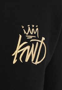 Guldbroderad stiliserad krona ovanför ordet "KNWLD" på svart tyg, som liknar graffitistil eller gatukonst.