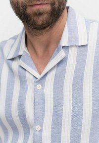 Chemise rayée bleu clair et blanche en tissu texturé, avec un col court et trois boutons, présentant des rayures alternant entre larges et étroites.