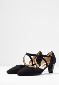 Scarpe con tacco alto in suede nero, caratterizzate da punta affusolata e cinturino incrociato alla caviglia con fibbia. Il tacco basso e bloccato offre stabilità.