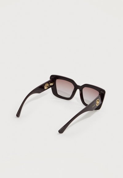 Μαύρα γυαλιά ηλίου με τετράγωνο πλαίσιο, φακούς με βαθμιαία απόχρωση και χρυσό μετάλλιο διακοσμητικό στα πλαϊνά τους, σε απλό ανοιχτό φόντο.