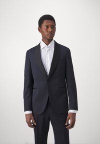 Marineblaues Formalsakko mit schwarzen Satinkleppen, Ein-Knopf-Verschluss und Taschen. Getragen über einem weißen Hemd.